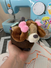 Load image into Gallery viewer, A brown yak plush. She has a furry dark brown coat and four grey hooves. She has a dark brown fluffy tail. She wears a green blanket on her back. She has a lighter brown face with pointed brown ears. She has grey horns. She has darker hair that is worn in two brown loops. She has two pink bows on her hair. Her muzzle is cream with a grey nose shaped like an oval. Her eyes are green.