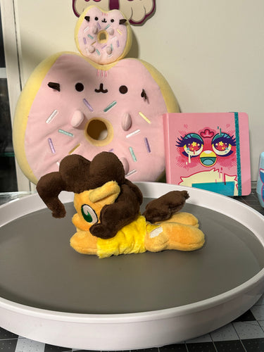 Yellow horse plush laying on belly with sandwich that has cheese in an arc between the slices on flank and brown hair. Eye is green. Wearing a yellow shirt. Plush is on my desk that has various pink decorations on it.