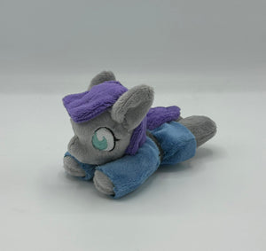 Grey horse plush laying on belly with purple hair. Eye is blue. Wears a blue dress with a grey belt