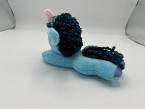 Blue horse plush laying on belly with blue yarn hair. Eye is green. Has a pink horn 