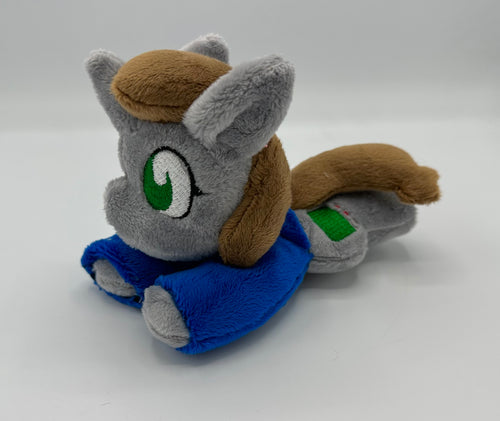 Grey horse plush laying on belly with green square on flank and two brown hair. Eye is green. Wears a blue shirt.