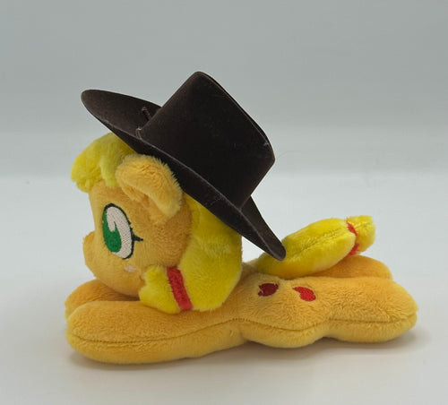 Orange horse plush laying on belly with red apples on flank and yellow hair with red hair ribbons. Eye is green. Wears a brown hat.