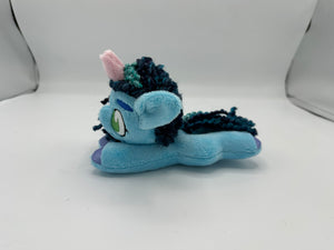 Blue horse plush laying on belly with blue yarn hair. Eye is green. Has a pink horn 