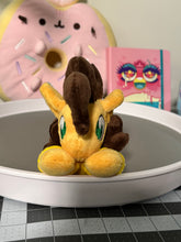 Load image into Gallery viewer, Yellow horse plush laying on belly with sandwich that has cheese in an arc between the slices on flank and brown hair. Eye is green. Wearing a yellow shirt. Plush is on my desk that has various pink decorations on it.
