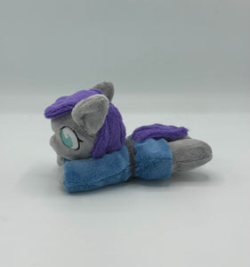 Grey horse plush laying on belly with purple hair. Eye is blue. Wears a blue dress with a grey belt