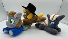 Load image into Gallery viewer, Grey horse plush laying on belly with green square on flank and two brown hair. Eye is green. Wears a blue shirt. She is joined by two of her friends. One is brown with a black hat and the other is grey with white hair. 
