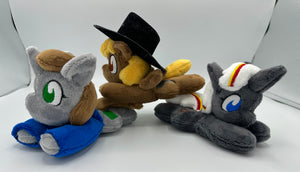 Grey horse plush laying on belly with green square on flank and two brown hair. Eye is green. Wears a blue shirt. She is joined by two of her friends. One is brown with a black hat and the other is grey with white hair. 