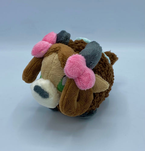 A brown yak plush. She has a furry dark brown coat and four grey hooves. She has a dark brown fluffy tail. She wears a green blanket on her back. She has a lighter brown face with pointed brown ears. She has grey horns. She has darker hair that is worn in two brown loops. She has two pink bows on her hair. Her muzzle is cream with a grey nose shaped like an oval. Her eyes are green. 