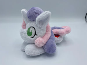 White horse plush laying on belly with purple and pink crest on flank and purple and pink hair. Eye is green.