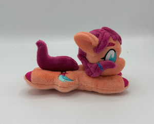 Coral colored horse plush laying on belly with pink stars and a blue trail on flank and maroon hair with blue hair tie. Eye is blue.