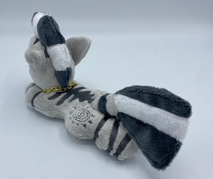 A zebra plush laying on belly. The coat is grey with black stripes. There is a grey sun design on the flank. She has a dark grey muzzle. Hair is white and grey stripes. Eye is blue. She has a gold chain around her neck. 