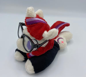 Cream colored horse plush laying on belly with purple moon and pink stars on flank and red hair with purple stripes. Eye is purple. Wears a black shirt and glasses