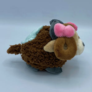 A brown yak plush. She has a furry dark brown coat and four grey hooves. She has a dark brown fluffy tail. She wears a green blanket on her back. She has a lighter brown face with pointed brown ears. She has grey horns. She has darker hair that is worn in two brown loops. She has two pink bows on her hair. Her muzzle is cream with a grey nose shaped like an oval. Her eyes are green. 