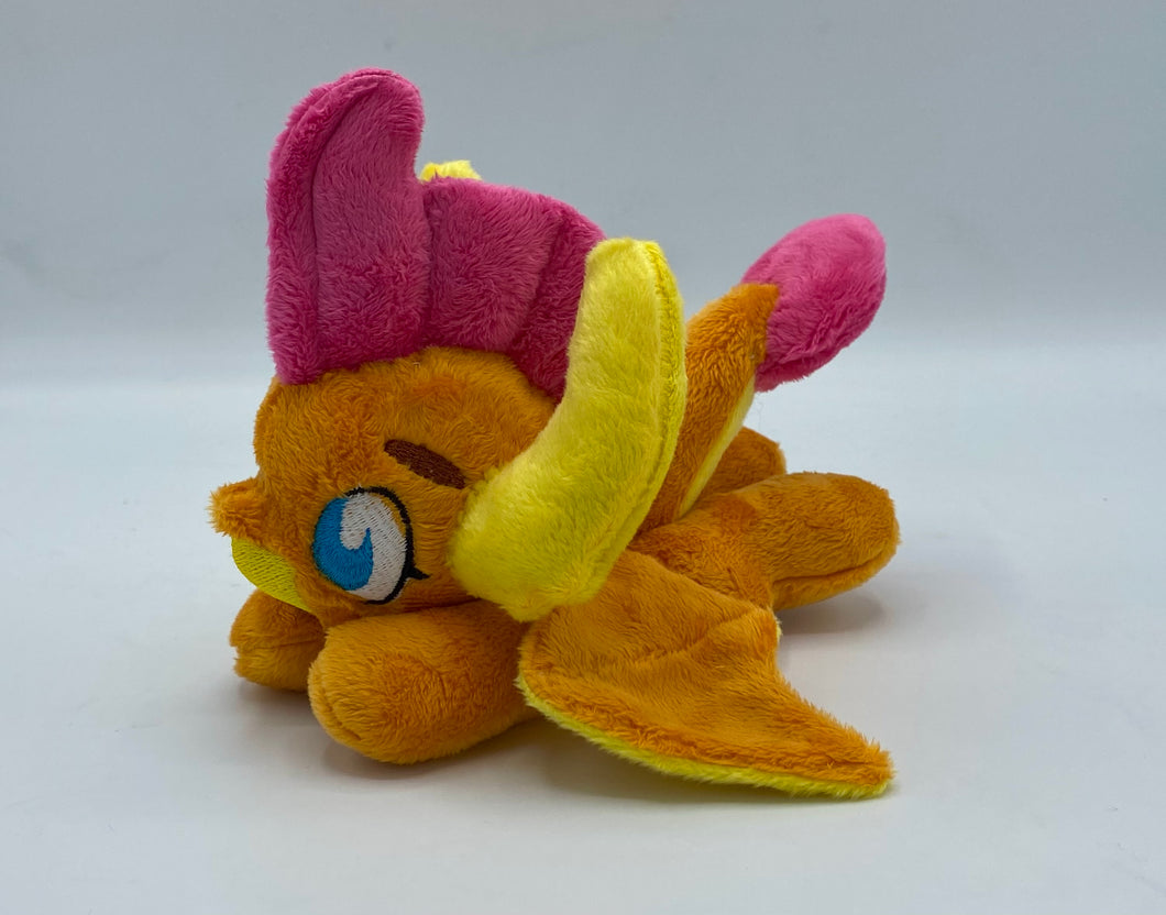 Orange dragon plush laying on belly. She is orange with a yellow belly. Her wings are orange and yellow. Her horns are yellow. She has pink hair. Her tail is orange and yellow with a pink tip. Her mouth is yellow. Her eyes are blue. She has brown eyebrows. 