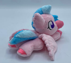 Pink hippogriff plush laying on belly. She has pink bird front feet and pink hooves on back feet. She has pink wings and a pink beak. Her eyes are purple. She has a pink ear that looks like feathers. Her hair is two tone blue. 