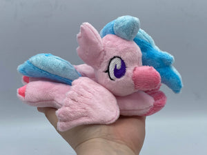 Pink hippogriff plush laying on belly. She has pink bird front feet and pink hooves on back feet. She has pink wings and a pink beak. Her eyes are purple. She has a pink ear that looks like feathers. Her hair is two tone blue. 