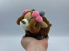 Load image into Gallery viewer, A brown yak plush. She has a furry dark brown coat and four grey hooves. She has a dark brown fluffy tail. She wears a green blanket on her back. She has a lighter brown face with pointed brown ears. She has grey horns. She has darker hair that is worn in two brown loops. She has two pink bows on her hair. Her muzzle is cream with a grey nose shaped like an oval. Her eyes are green. 
