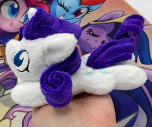 White horse plush laying on belly with blue diamonds on flank and purple curly hair. Eye is blue.