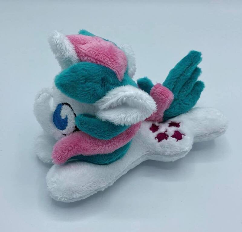 White horse plush laying on belly with purple leaves on flank and pink and green hair. Eye is blue. Has pink bow on tail